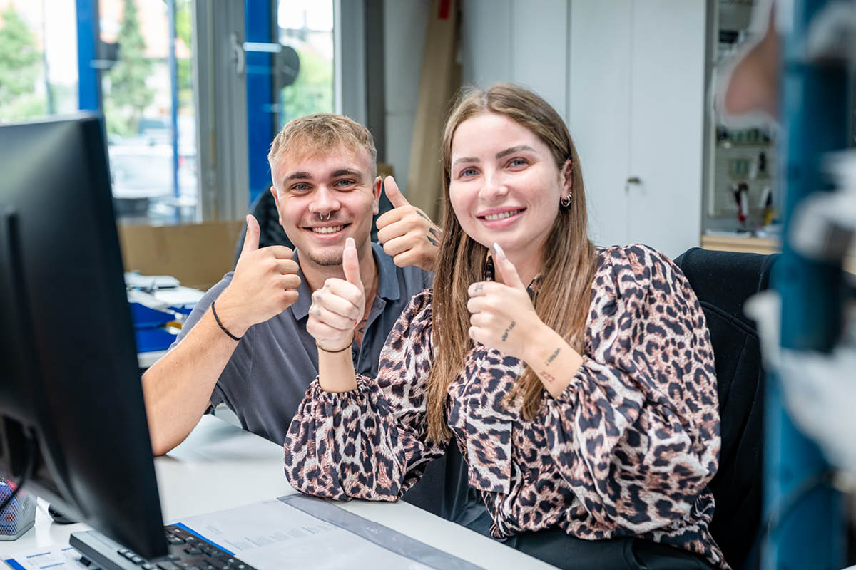Zwei Mitarbeiter sitzen im Büro und zeigen beide Daumen nach oben in die Kamera