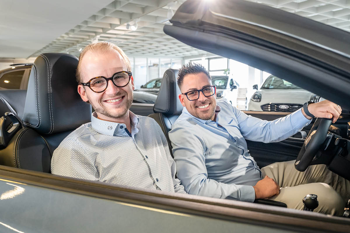 Zwei Mitarbeiter sitzen in einem Auto und lachen in die Kamera